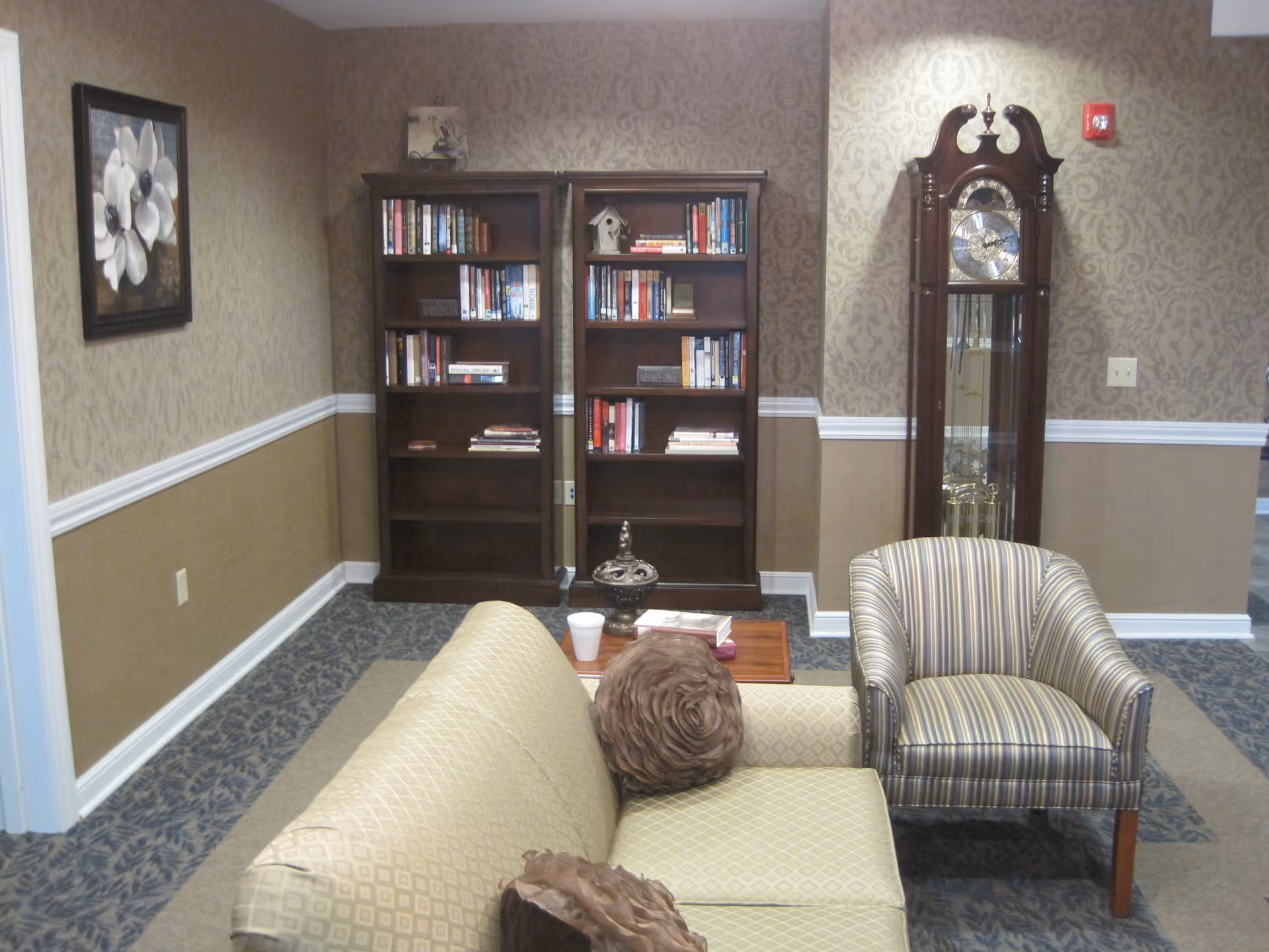 A cozy living room area with a beige patterned sofa adorned with two decorative pillows, a striped armchair, a wooden coffee table with a cup and books, two dark wooden bookshelves filled with books, a tall grandfather clock, and a framed floral painting on the wall. The walls have a two-tone design with wallpaper on the upper half and a solid color on the lower half, separated by white chair rail molding.