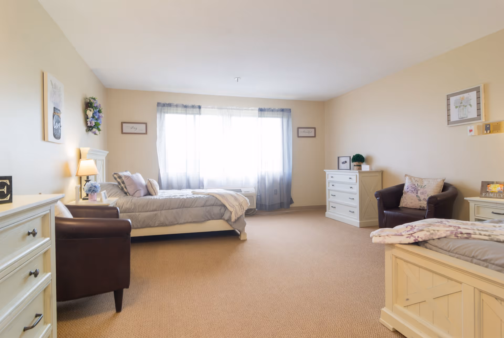 A spacious, well-lit bedroom with two beds, beige walls, and carpeted floor. The room features a large window with sheer curtains allowing natural light to fill the space. There are two armchairs, a dresser, and decorative wall art including framed pictures and a floral wreath.
