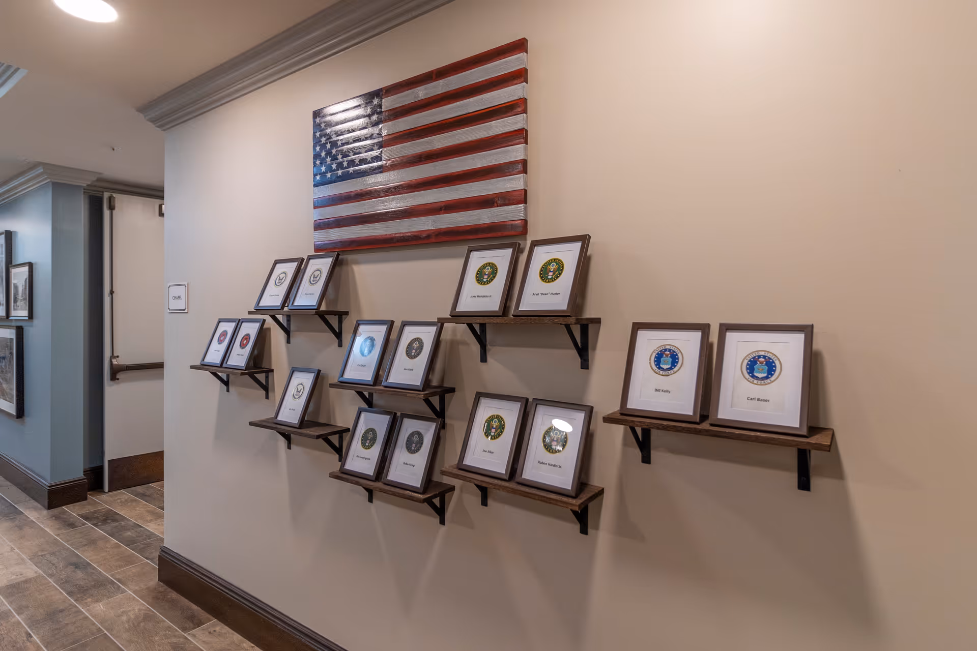 Interior hallway wall display with multiple framed certificates or plaques arranged on wooden shelves beneath a wooden American flag decoration. The wall is beige and the floor has brown tiles. A sign on the left side indicates a chapel nearby.