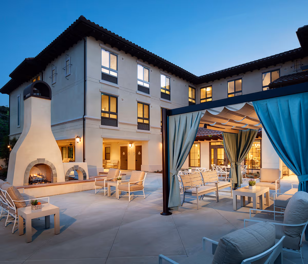 Outdoor patio area at Belmont Village Senior Living Calabasas featuring a large fireplace, multiple seating arrangements with cushioned chairs and benches, and a pergola with blue curtains. The building exterior is visible in the background with warm lighting from windows and wall-mounted lamps.