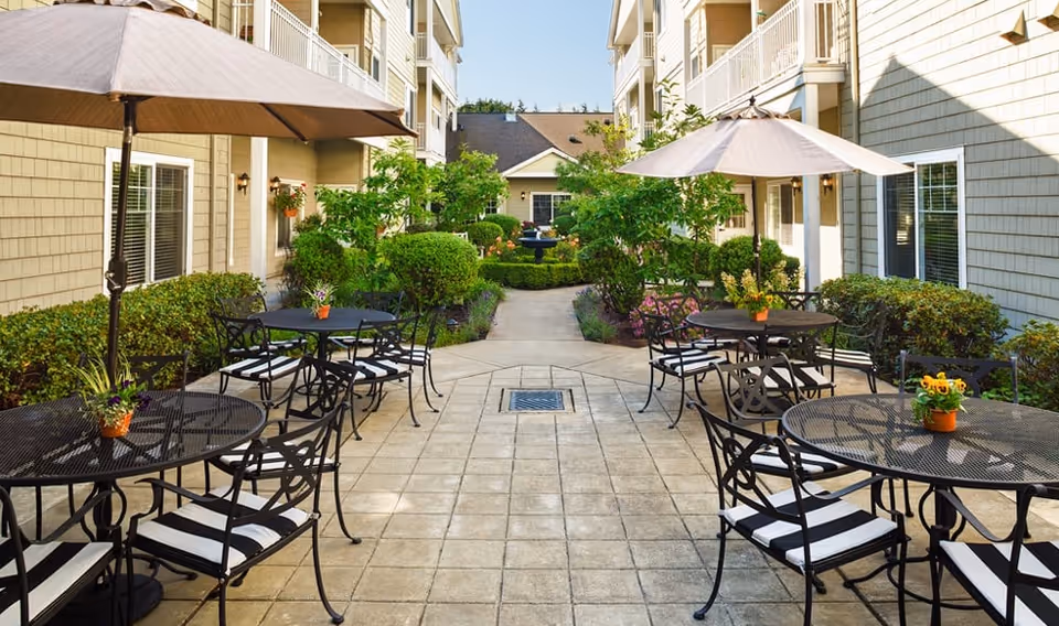 Outdoor patio area at Canterbury Park Independent Living Community with black metal tables and chairs featuring striped cushions, large umbrellas providing shade, potted plants on tables, and well-maintained greenery and bushes along the walkway between two buildings.