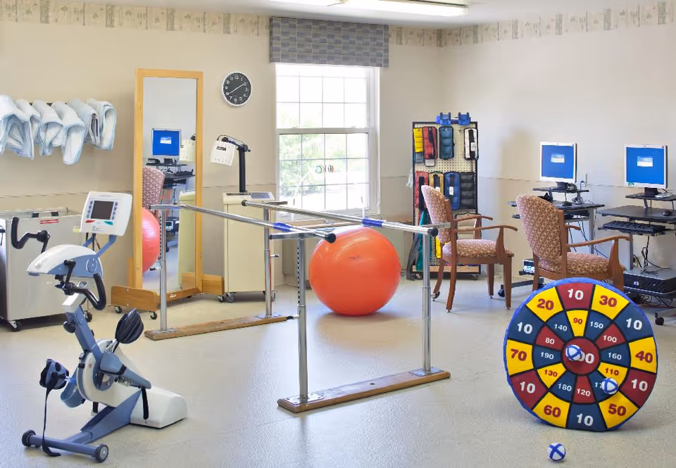 A physical therapy or rehabilitation room with exercise equipment including a stationary bike, parallel bars, a large red exercise ball, and a colorful target game with balls. There are two chairs in front of computer stations, a full-length mirror, and a rack with various exercise bands. A window lets in natural light.