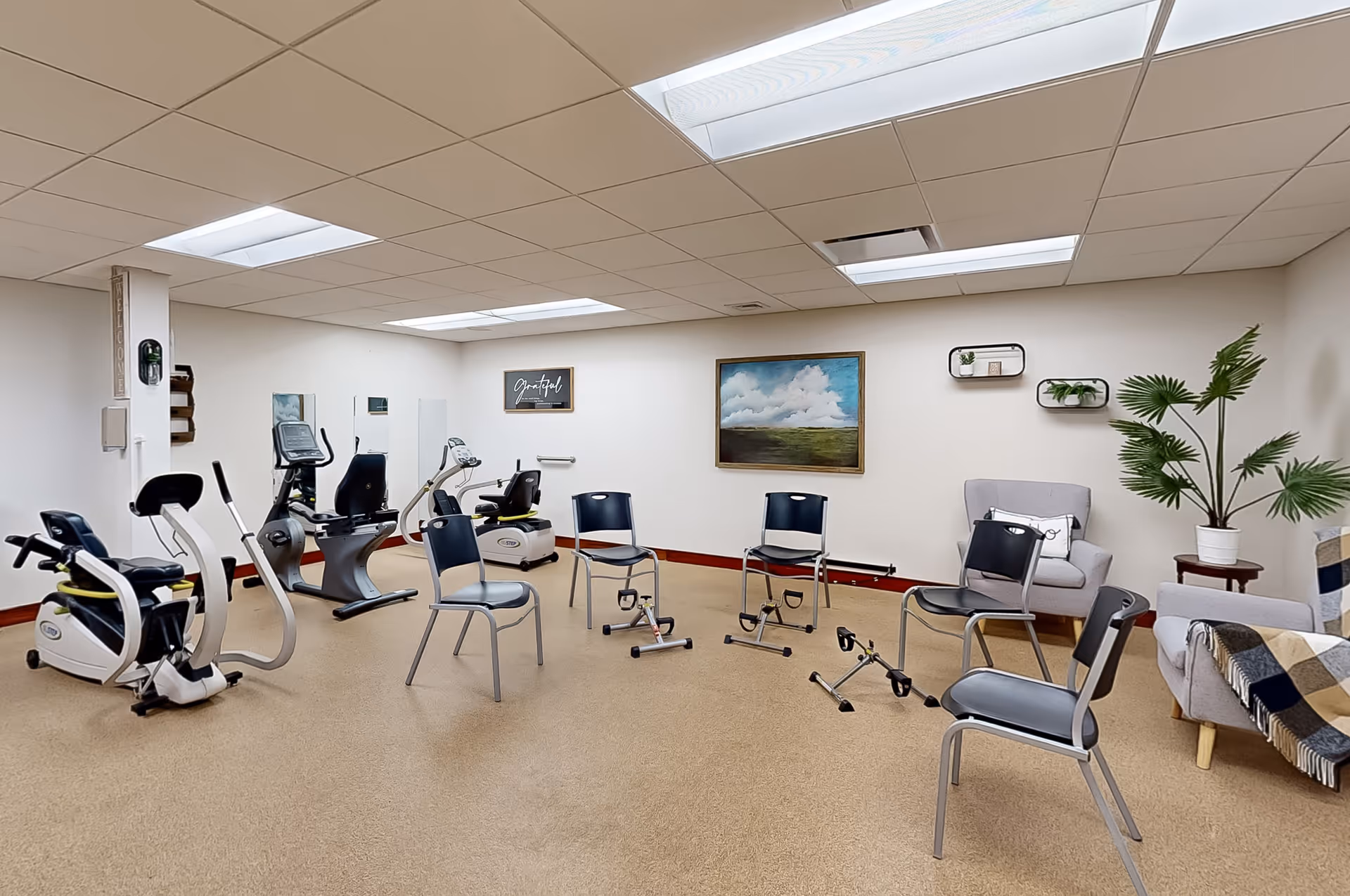 A well-lit exercise room with several stationary exercise bikes and chairs arranged in a circle. The room has beige carpet flooring, white walls, and a drop ceiling with fluorescent lights. There are framed artworks and small shelves with plants on the walls, along with a couple of armchairs and a small table with a potted plant in the corner.