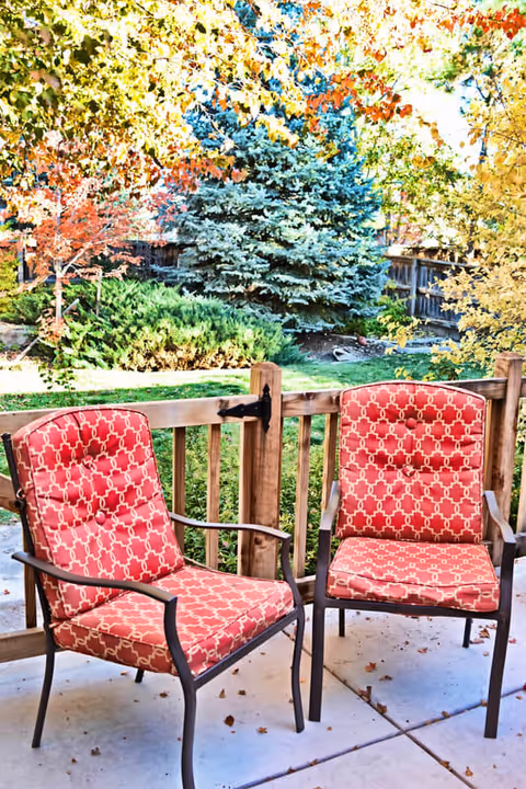 Two red cushioned patio chairs with a geometric pattern are placed on a concrete patio. Behind the chairs is a wooden fence, and beyond that is a garden area with green grass, bushes, and trees with autumn-colored leaves.