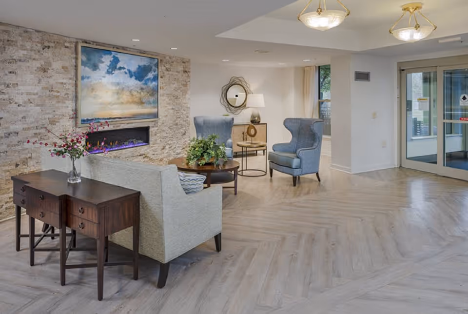 A cozy assisted living facility common area with a light-colored sofa, two blue armchairs, a wooden side table with a vase of flowers, a round coffee table with a plant, a modern electric fireplace set in a stone wall, and a large painting above the fireplace. The room has light wood flooring, two ceiling light fixtures, a round mirror on the wall, and glass doors leading outside.
