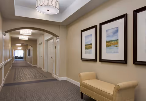 Well-lit carpeted hallway with framed artwork, a bench, and arched doorways in a senior living facility.