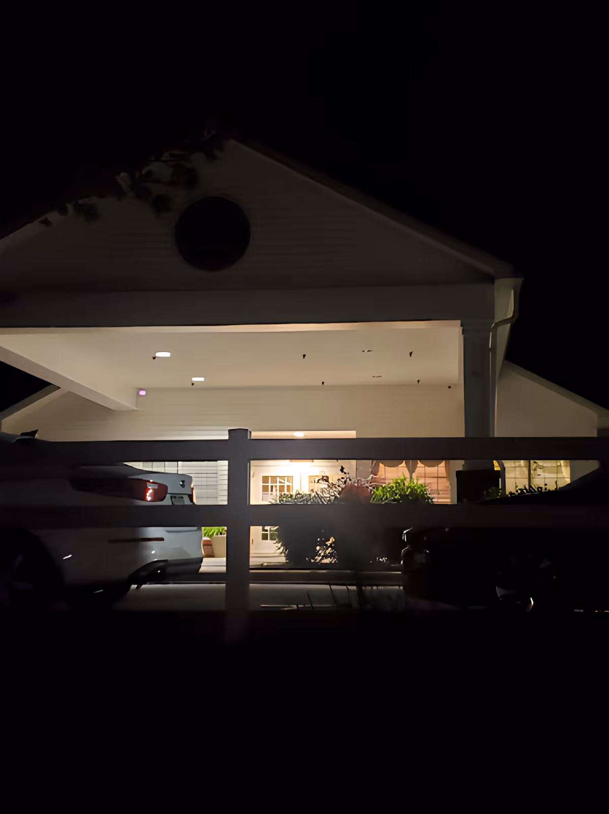Night view of the entrance area of a building with a white fence in the foreground. The building has a triangular roof with a circular window near the top. There are two cars parked on either side of the entrance, and plants are visible near the doorway, which is illuminated by warm lighting.