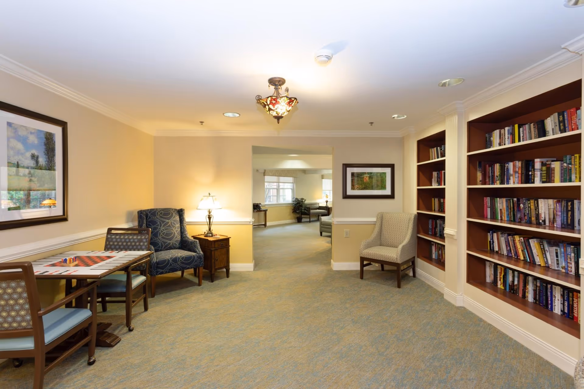 Communal lounge area with bookshelves, armchairs, a game table, and soft lighting leading to another seating area.