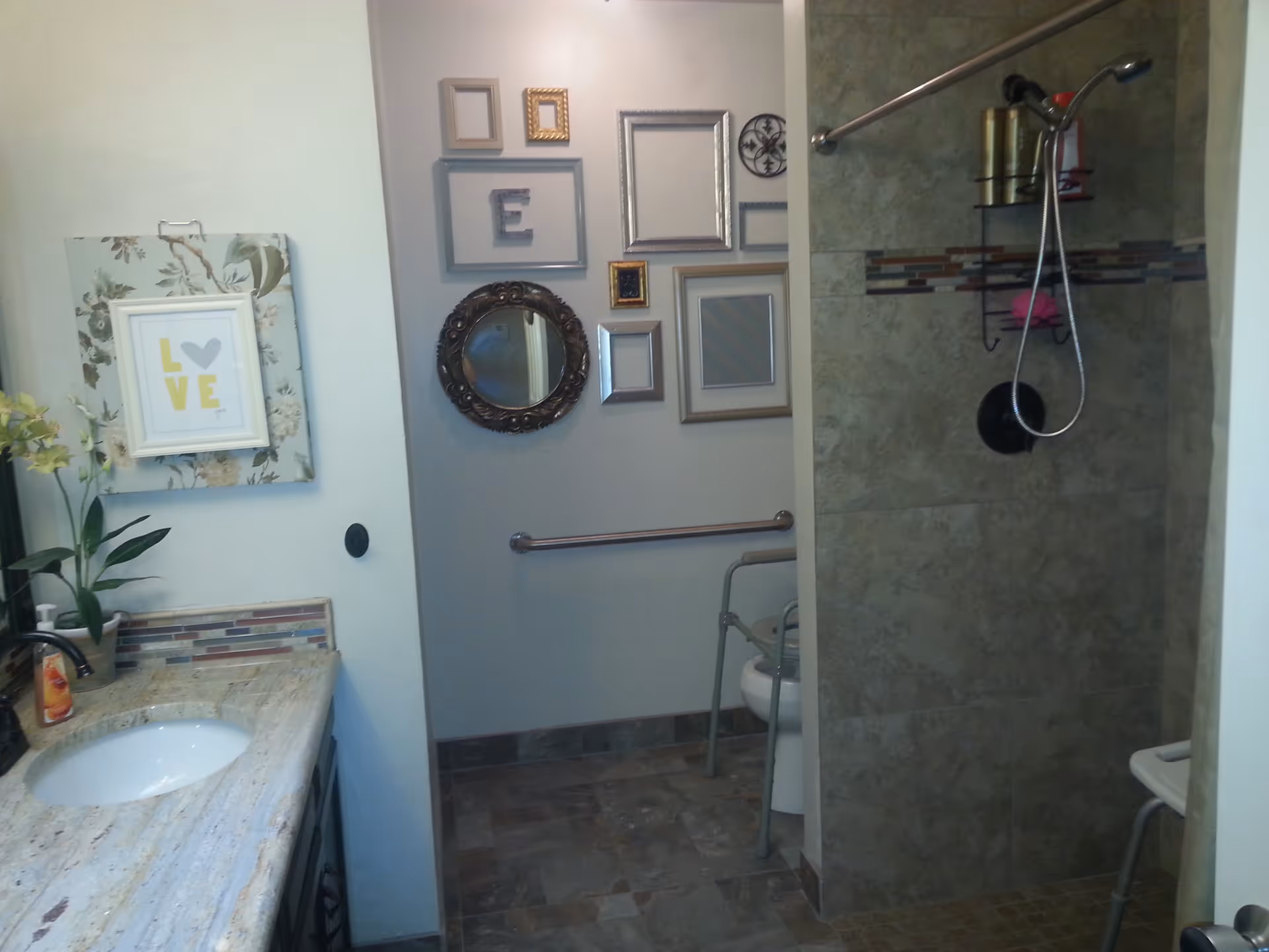 A bathroom with a granite countertop sink on the left, decorated with a small plant and a framed picture that says 'LOVE'. The back wall features multiple empty picture frames and a round mirror. On the right, there is a tiled walk-in shower with a shower chair and a handheld showerhead. A grab bar is mounted on the wall near the toilet, which also has a shower chair.