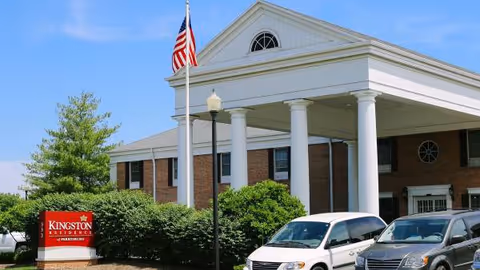 Front view of Kingston Residence Perrysburg, an assisted living building with columns, flag, and parked cars.