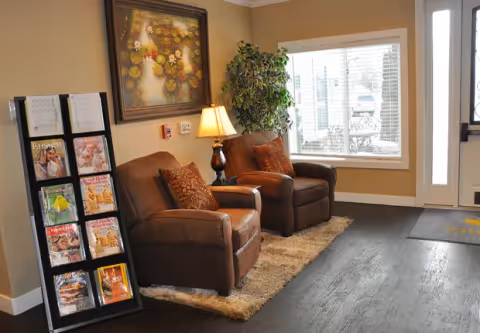 A cozy seating area in a senior living facility with two brown leather armchairs on a beige rug, a side table with a lamp, a large framed painting on the wall, a tall green plant near a window with white blinds, and a magazine rack filled with various magazines.