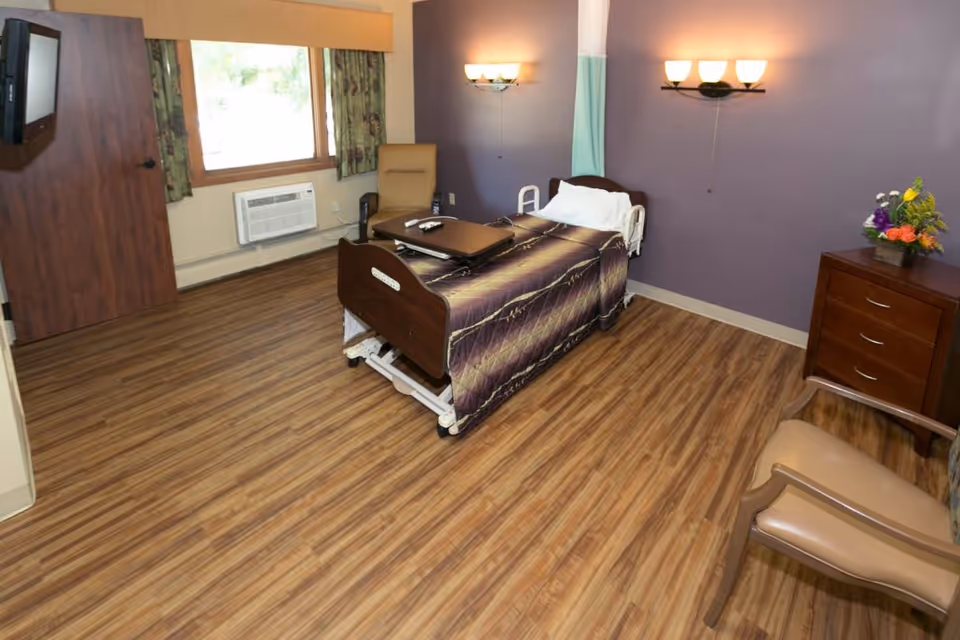A senior living resident room with a single adjustable bed, wood-look flooring, purple accent wall, chair, dresser with flowers, wall lights and a window with an air conditioner.