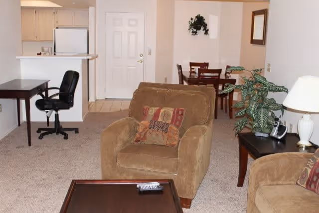 Open-plan living room showing upholstered armchairs and coffee table, side table with lamp, a small desk and chair, and a dining table near the kitchen and front door.