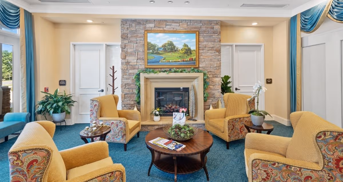 Cozy lounge with a stone fireplace, a framed painting above it, four upholstered armchairs around a central coffee table, and potted plants.