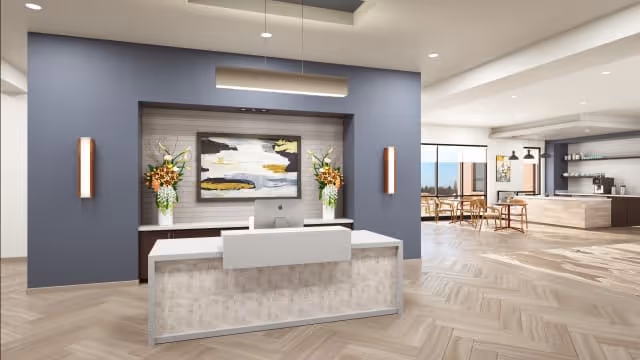 A modern reception area in a senior living facility with a front desk, two floral arrangements on either side, a large abstract painting on the wall behind the desk, and a spacious open area with tables and chairs in the background.