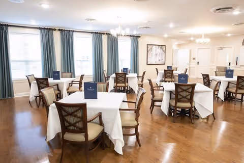 A bright dining room with several square tables covered with white tablecloths, each surrounded by wooden chairs with cushioned seats. Blue menus or signs are placed on each table. The room has large windows with blue curtains, wooden flooring, and white walls with framed artwork. Ceiling lights provide ample illumination.