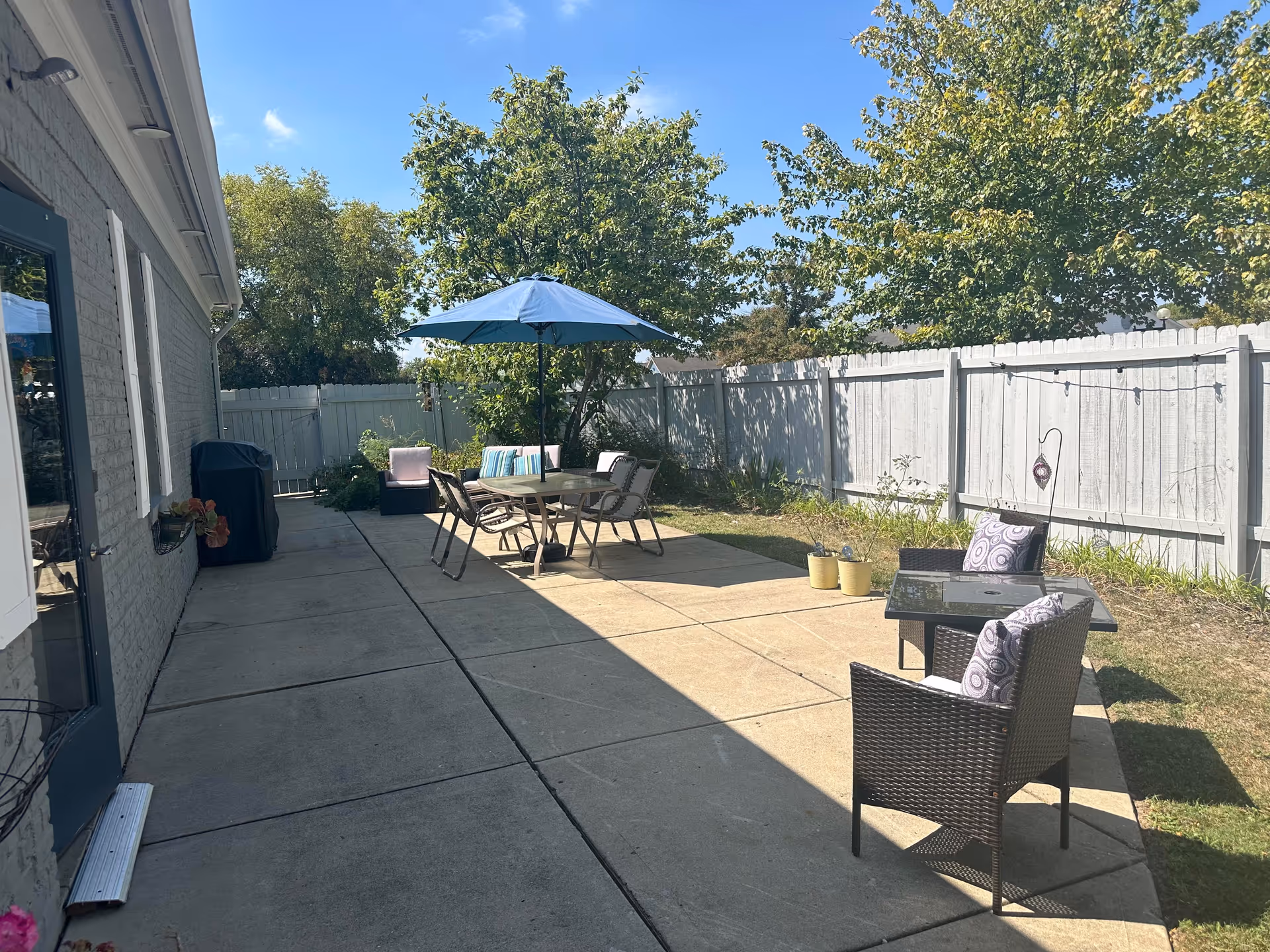 Sunny fenced patio area with an outdoor dining table under a blue umbrella, several chairs, and potted plants alongside a building.