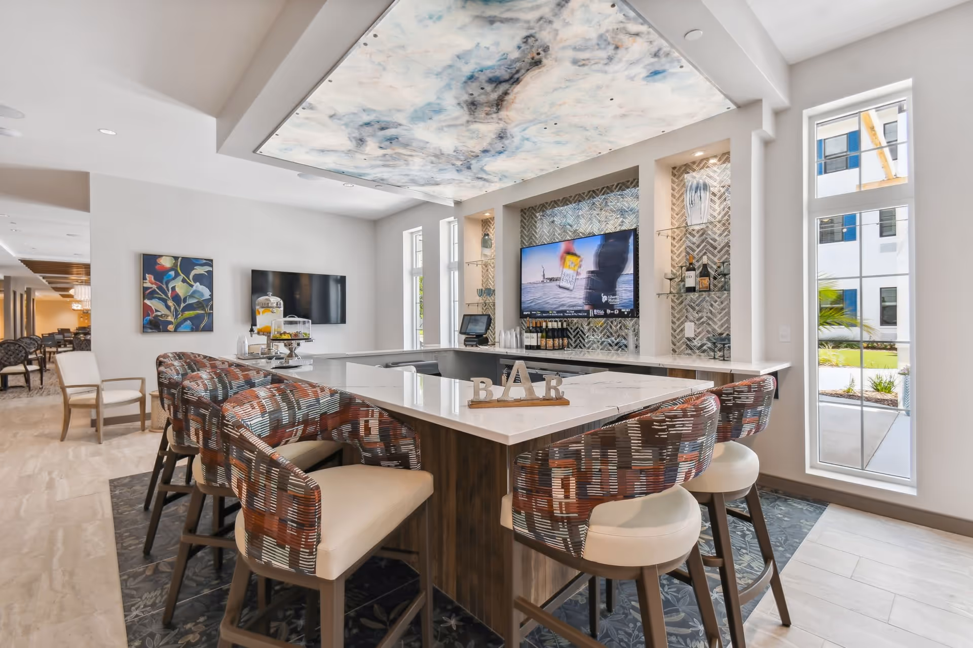 Modern senior living facility bar area with a white marble countertop, six patterned cushioned bar stools, a large TV mounted on a tiled wall, and a decorative ceiling panel with a cloudy sky design. Large windows allow natural light to brighten the space, and a small sign spelling 'BAR' is placed on the counter.