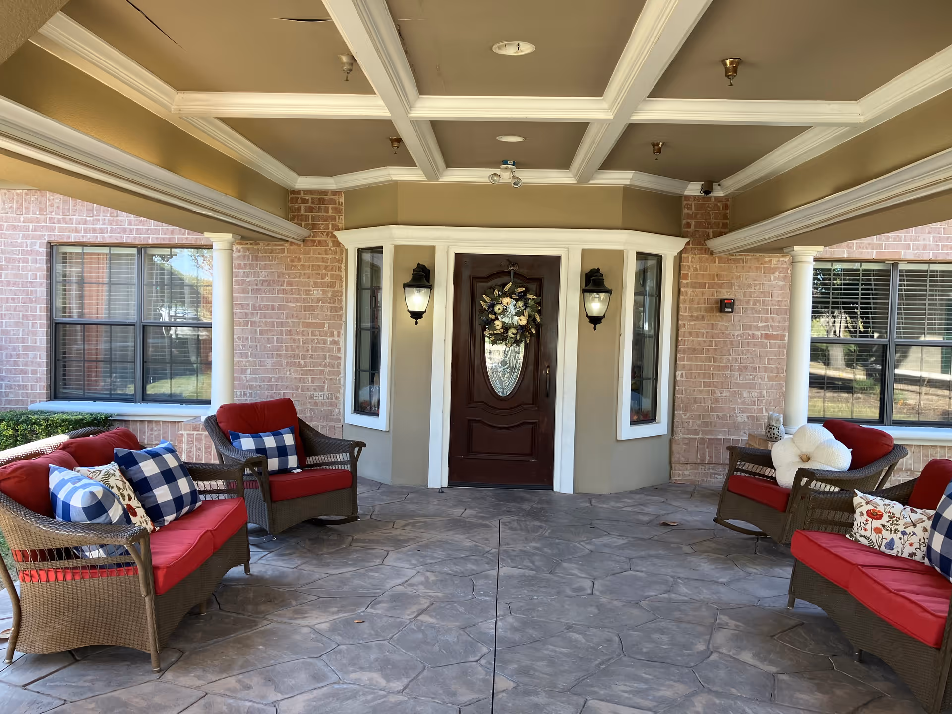 Covered front entrance porch with wicker seating and red cushions facing a decorative wood door with a wreath.