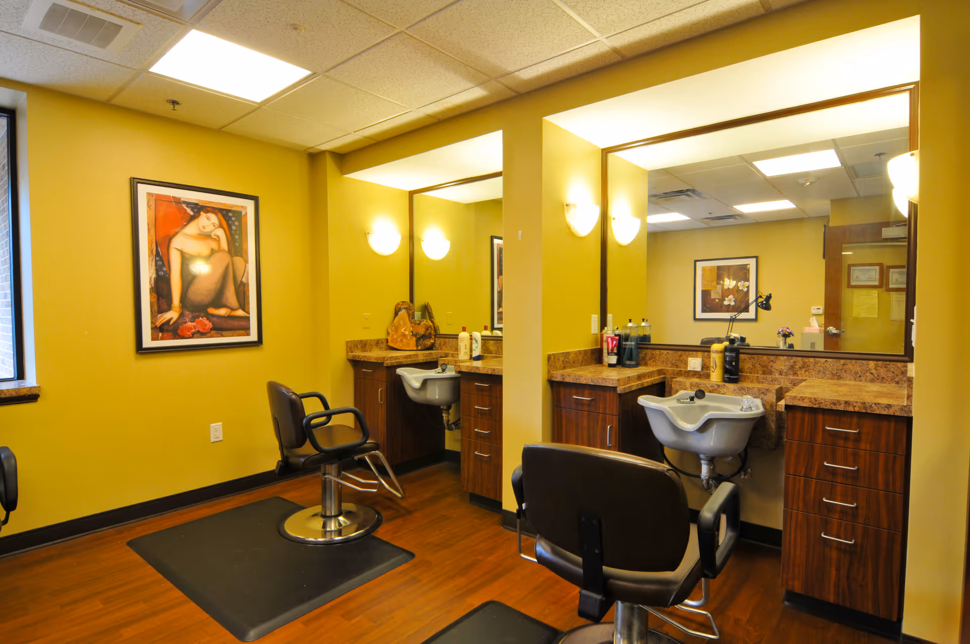 Interior view of a hair salon area with two salon chairs in front of large mirrors and sinks, wooden cabinets, yellow walls, and framed artwork on the walls.
