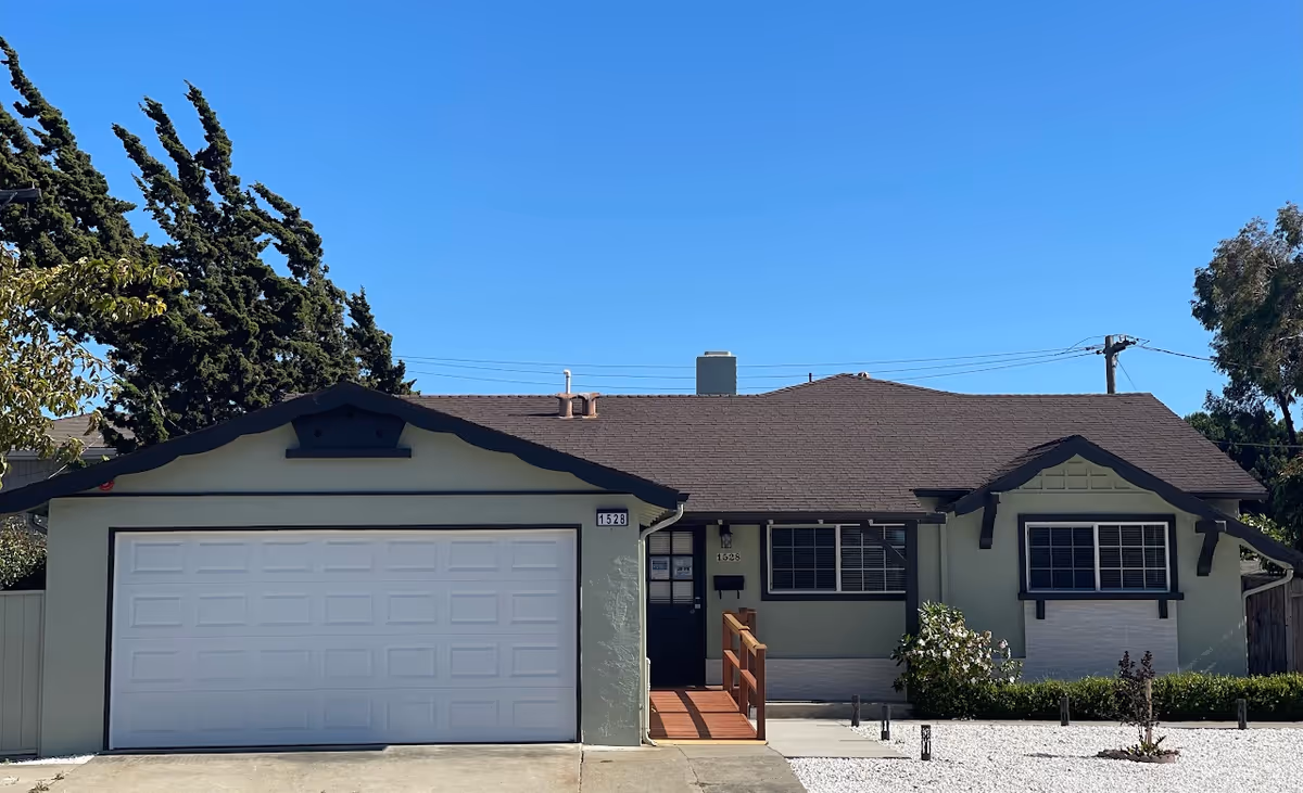 Single-story light-green house with an attached garage, small front porch ramp, and gravel front yard under a clear blue sky.