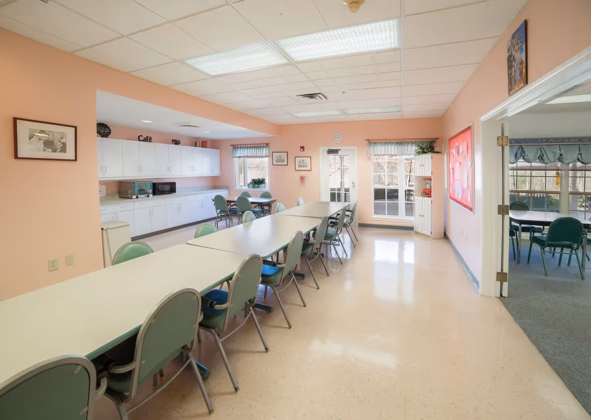 A bright dining room with long tables and green chairs arranged around them. The walls are painted peach, and there are white cabinets along one side with a microwave and toaster oven on the counter. Two windows with valances and a glass door lead to an outdoor area. A bulletin board and framed pictures decorate the walls.