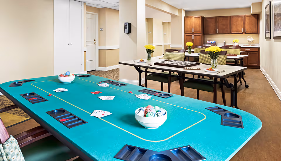 A recreational room in a senior living facility featuring a green poker table with playing cards and poker chips, and a table in the background with chairs, yellow flower vases, and a board game. The room has beige walls, wooden cabinets, and a hardwood floor.