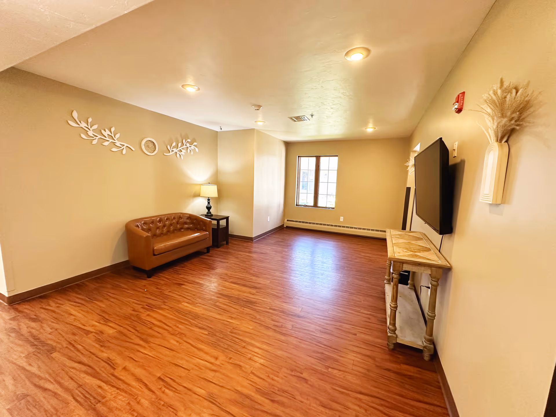 A spacious room with wooden flooring and beige walls featuring a brown leather loveseat, a small side table with a lamp, a wall-mounted flat screen TV above a wooden console table, and decorative wall art including white branches and a circular piece. A window allows natural light into the room.