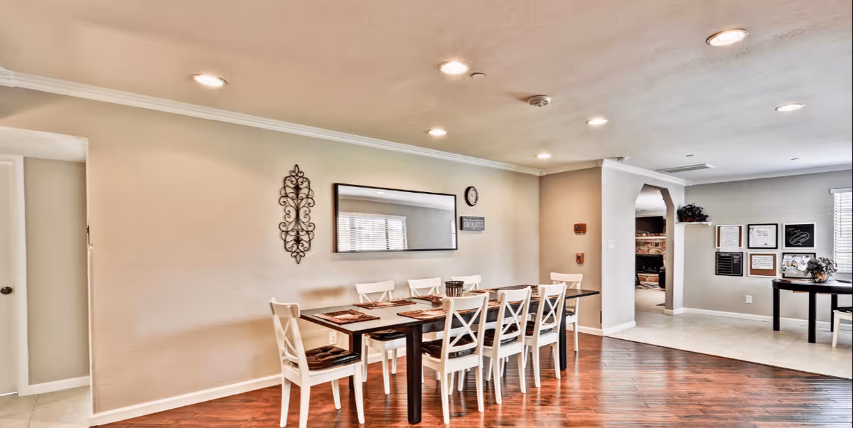 A bright dining area with a long dark wooden table surrounded by eight white chairs with dark cushions. The walls are beige with a decorative metal wall hanging, a large rectangular mirror, and a clock. The floor is a mix of dark wood and light tile, and recessed ceiling lights illuminate the space. In the background, there is an open doorway leading to another room with a fireplace and framed pictures on the walls.
