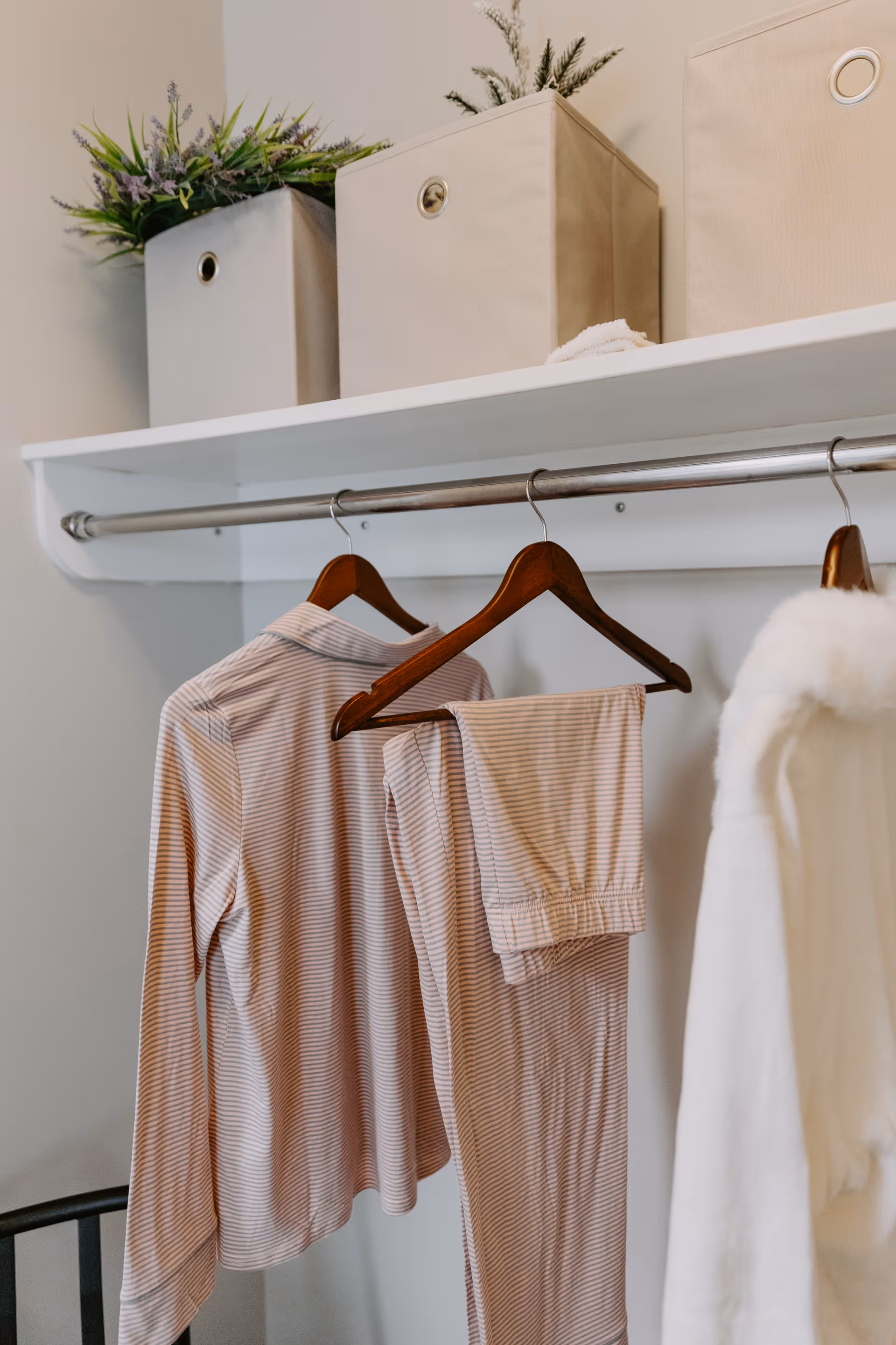 A closet rod with wooden hangers holding a striped pajama set and a white furry garment. Above the rod is a white shelf with three beige fabric storage bins, two of which have green plants inside.