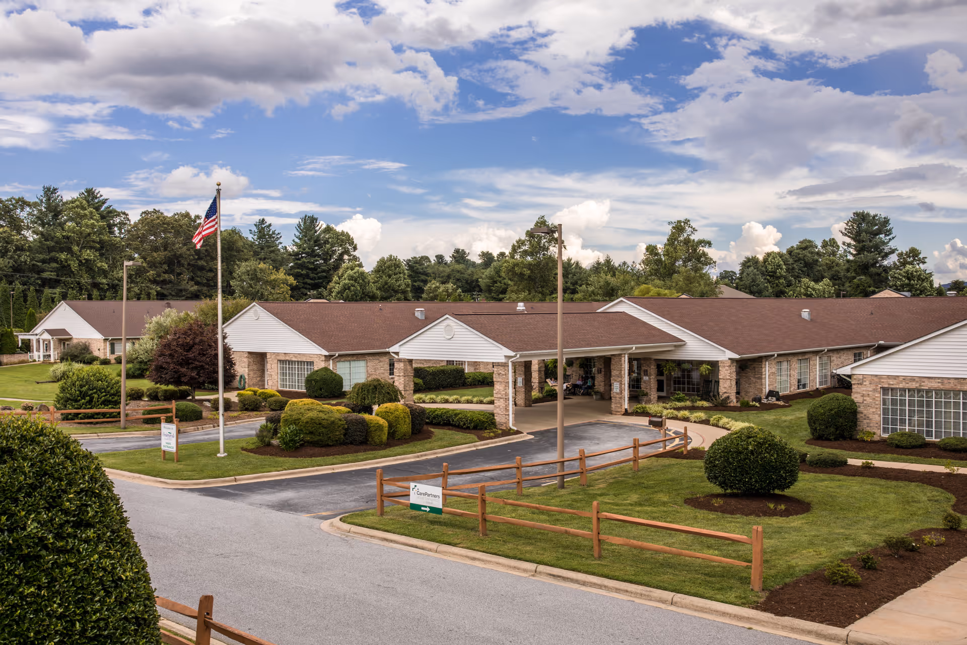Front exterior of a single-story senior living facility with a covered entrance, American flag, driveway, and manicured landscaping.