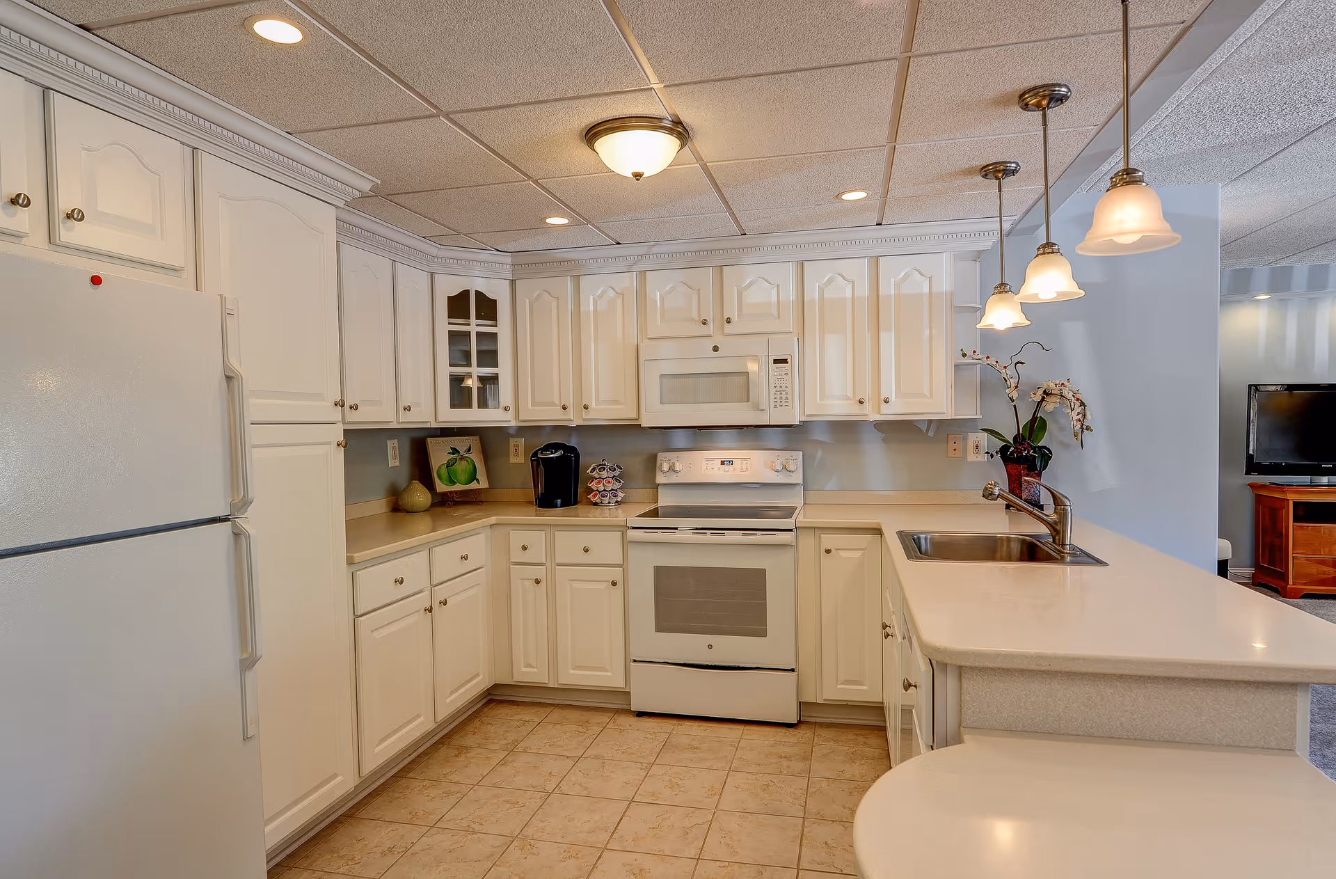 A clean, well-lit kitchen with white cabinets, a white refrigerator, a white stove with an overhead microwave, and a sink on a beige countertop. There are pendant lights hanging above the counter, a coffee maker, a decorative plant, and a small framed picture on the counter. In the background, part of a living room with a TV is visible.