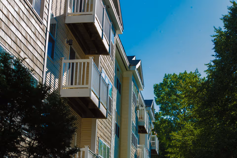 Exterior facade of a multi-story residential building showing balconies and trees against a blue sky.