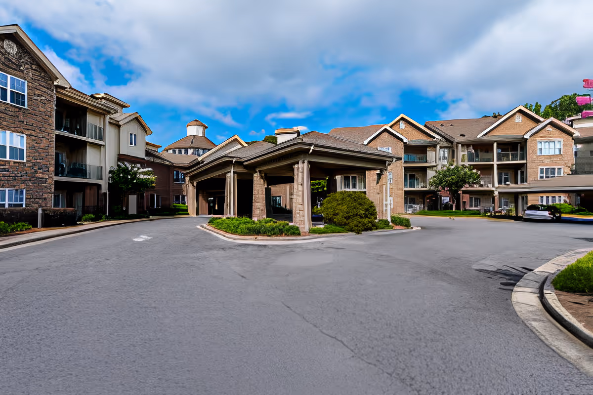 Exterior view of The Haven at Regency Pointe showing a multi-story senior living facility with a covered entrance driveway, surrounded by landscaped greenery under a partly cloudy blue sky.