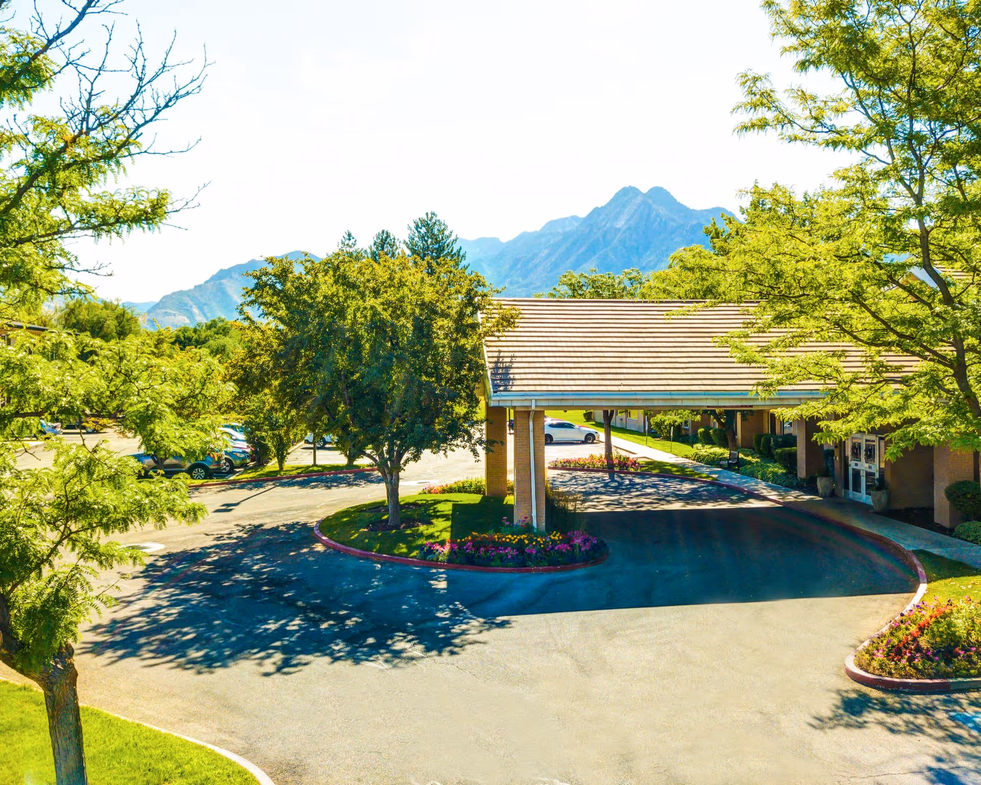 Exterior view of The Auberge at Aspen Park facility entrance with a covered driveway, surrounded by trees and landscaped flower beds, with mountains visible in the background under a clear sky.