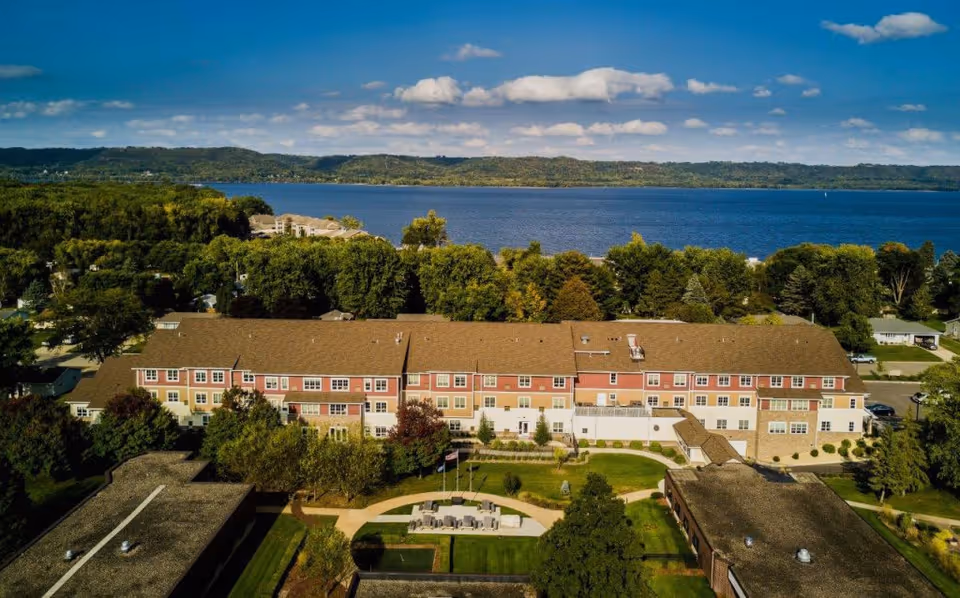Aerial view of a multi-story senior living building with courtyards, lawns and trees on the shore of a large lake.