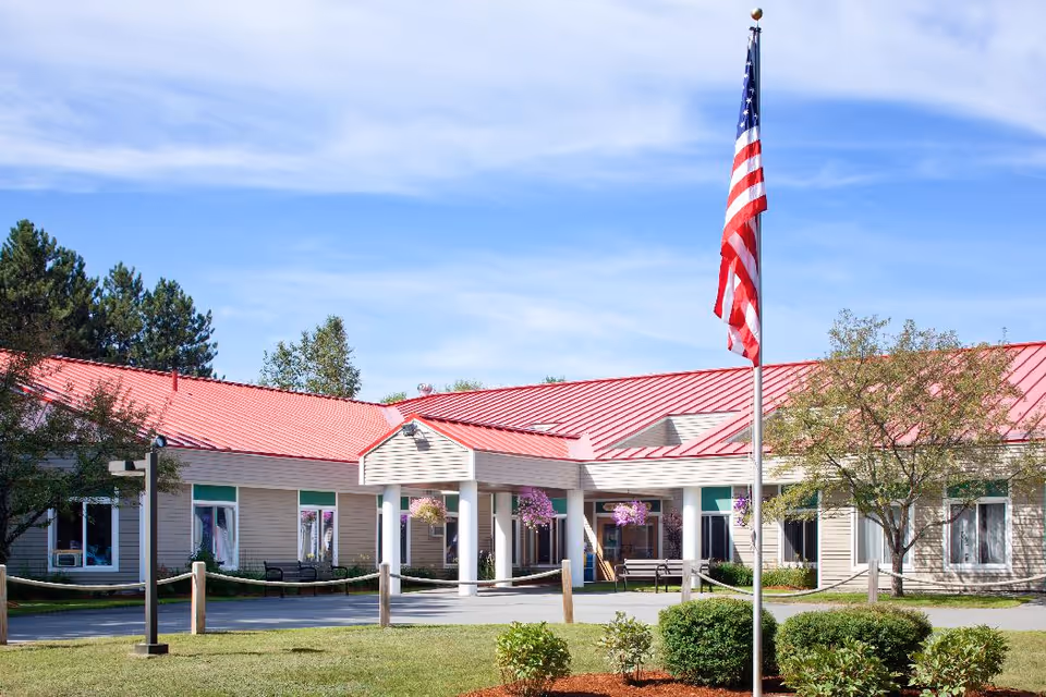Exterior view of Cedar Ridge Center, a single-story building with a red roof, beige siding, and white columns at the entrance. There is an American flag on a flagpole in front of the building, surrounded by green bushes and trees under a partly cloudy blue sky.