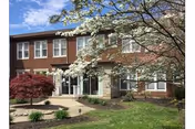 Exterior view of a two-story brick building with white-framed windows, surrounded by landscaped greenery including a small tree with white blossoms and a red-leafed shrub under a partly cloudy blue sky.