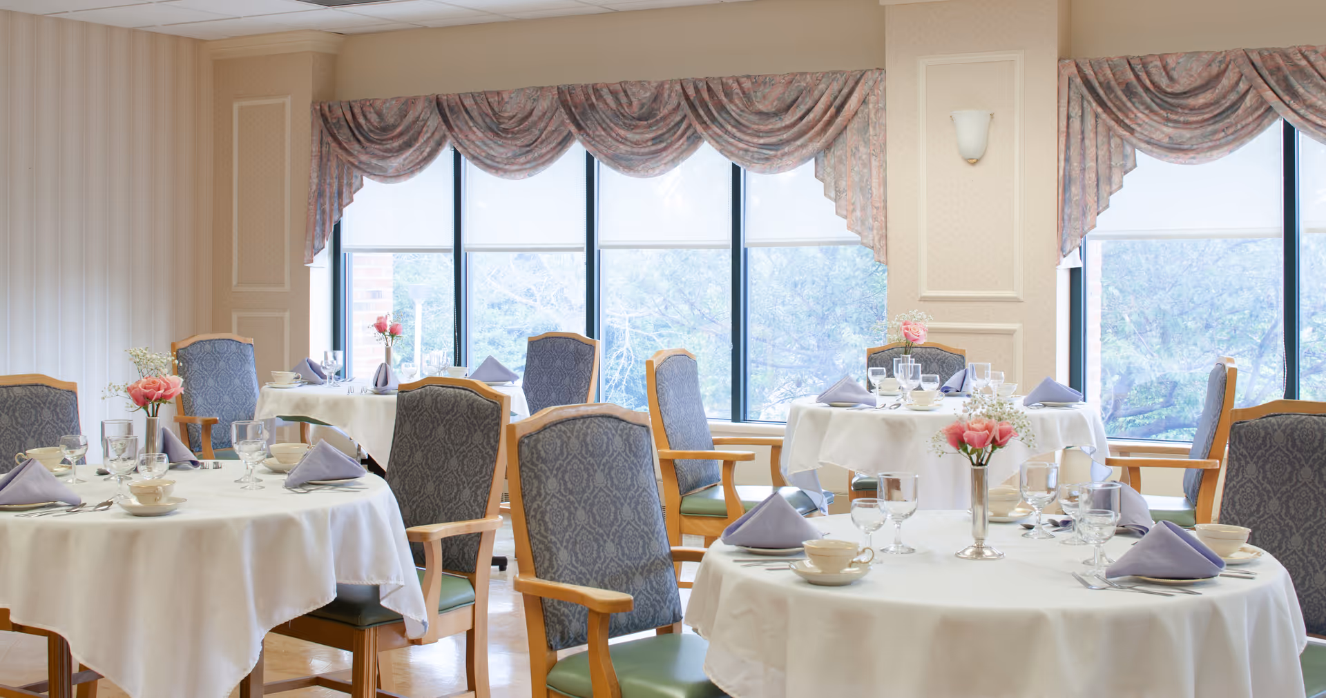 Bright dining room with round tables set with white tablecloths, folded napkins, glassware, and floral centerpieces by large windows.