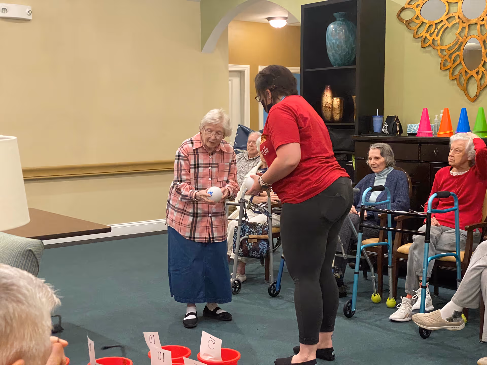 A staff member helps an elderly woman hold a ball during an activity as other residents watch in a common room.