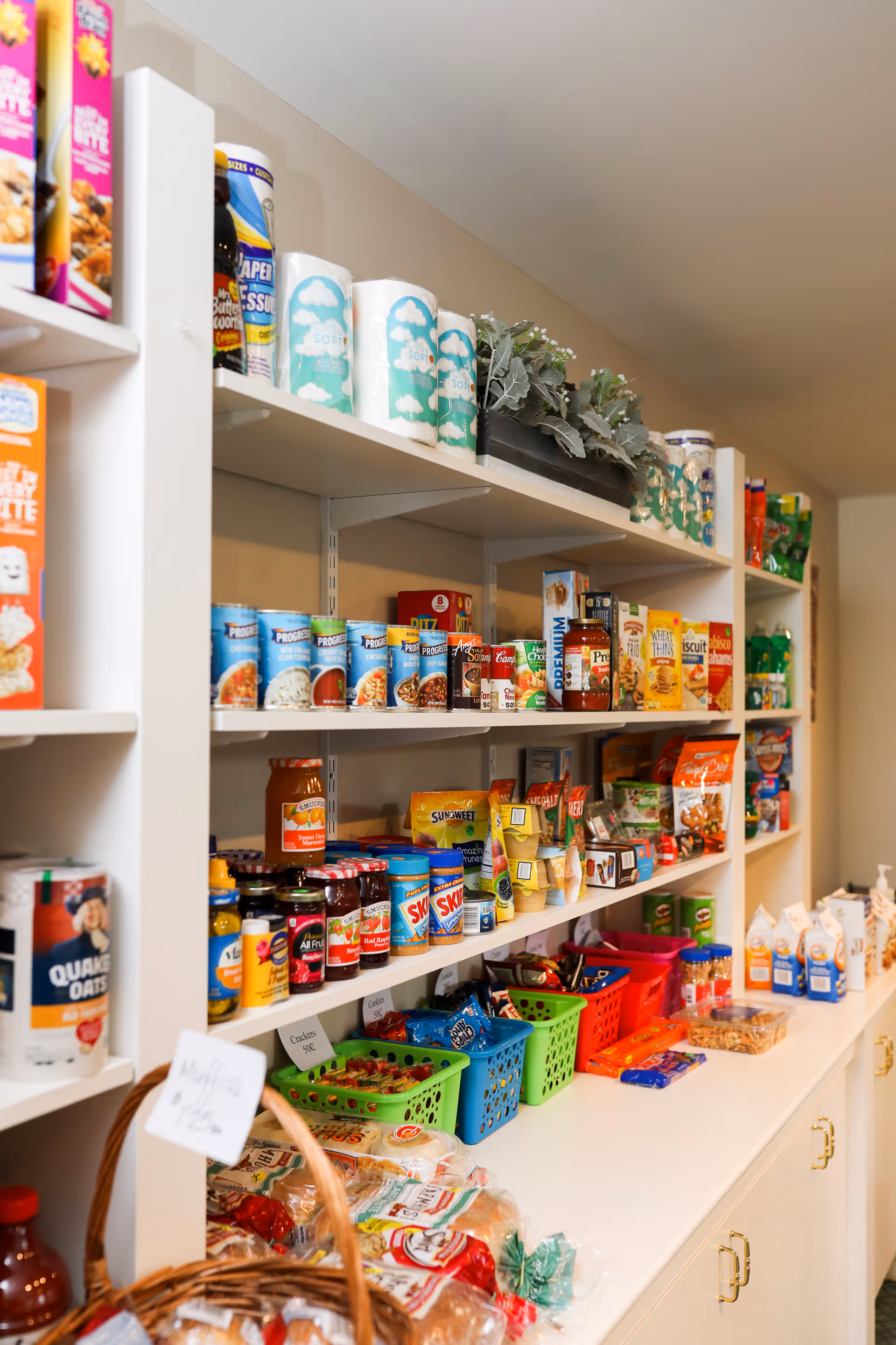 Shelves stocked with various food items including canned goods, cereals, snacks, and paper towels in a pantry or storage area. There are colorful plastic baskets holding snacks and other packaged foods on the counter below the shelves.