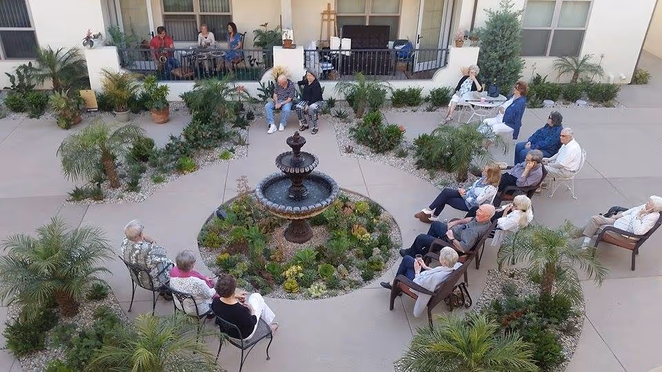 A group of elderly people sitting in a circular outdoor courtyard around a central water fountain surrounded by plants and small palm trees. Some people are seated on metal chairs, others on cushioned chairs, and a few are sitting on a raised porch area in the background.