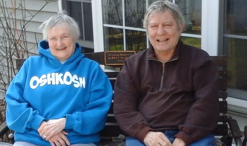 An elderly woman wearing a blue Oshkosh hoodie and an elderly man wearing a dark brown jacket sitting together on a wooden bench outside a building with windows behind them. There is a small plaque on the bench in memory of Karen J. Seeliger.