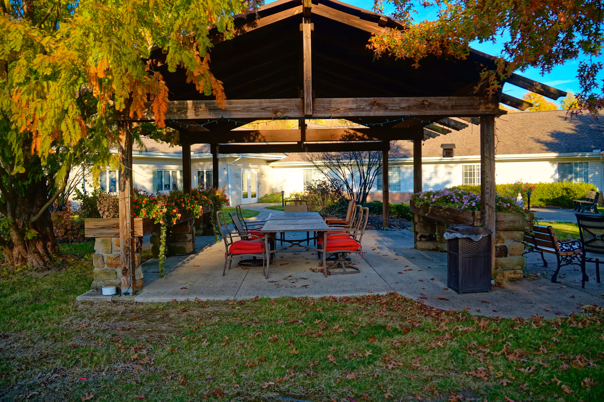 A covered outdoor pavilion with a table and red-cushioned chairs surrounded by planters and greenery in a senior living courtyard.