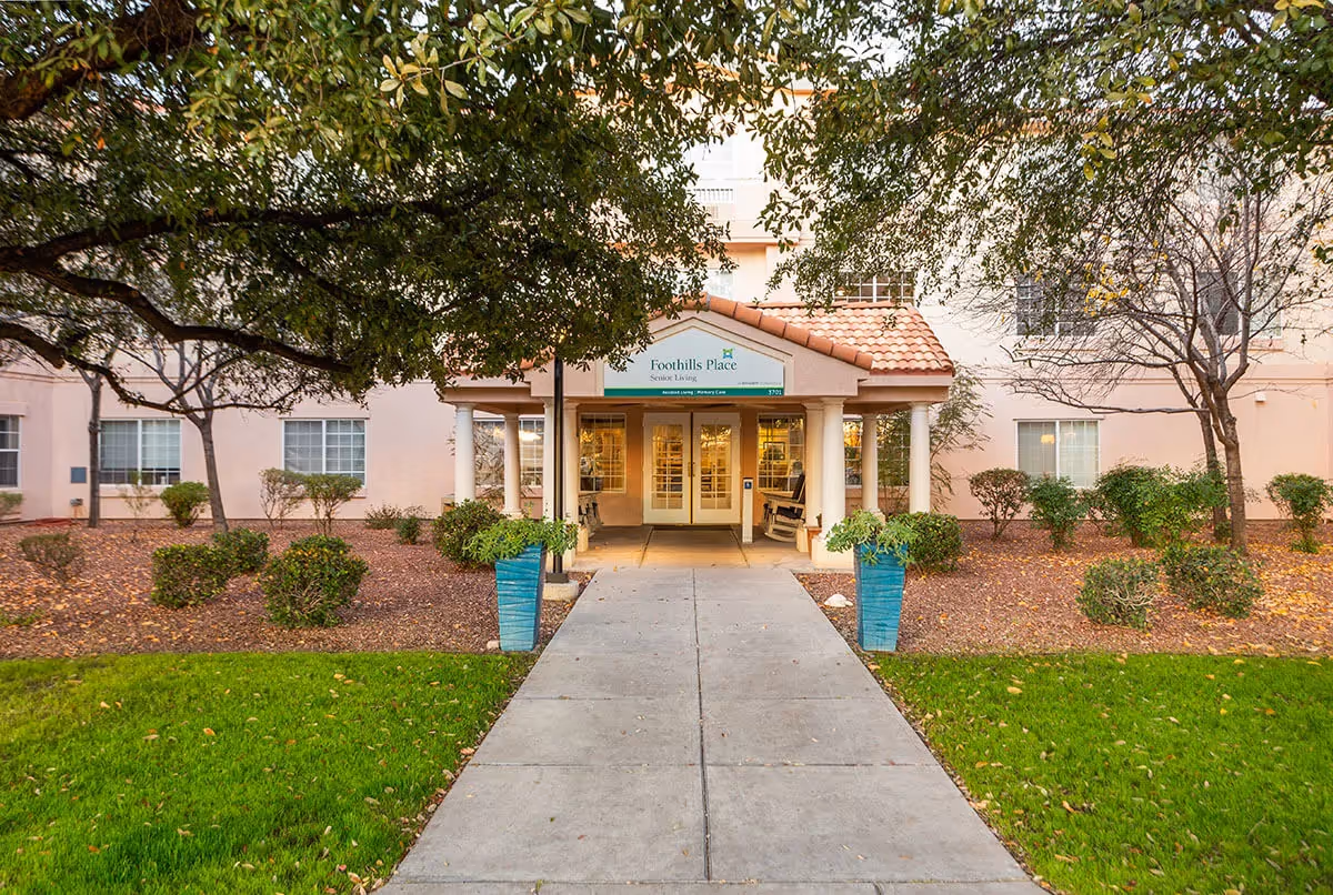 Entrance to a senior living facility named Foothills Place with a covered walkway supported by columns, flanked by two large blue planters with greenery. The building is light-colored with several windows, surrounded by landscaped bushes, trees, and a green lawn.
