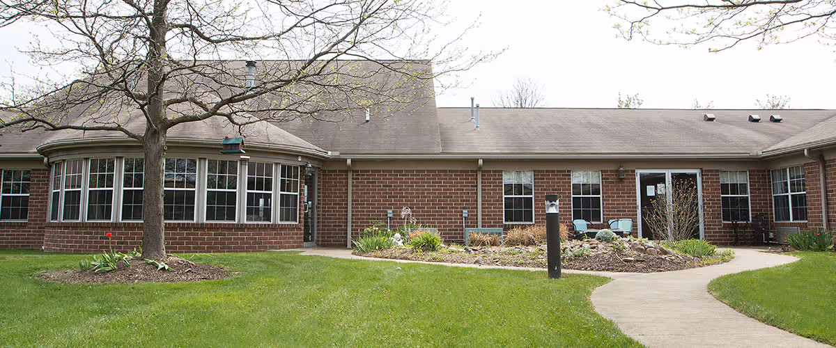 Exterior view of a single-story brick building with multiple windows and a curved section on the left side. There is a leafless tree in front of the building, a well-maintained lawn, a garden bed with plants, and a curved concrete walkway leading to the entrance.