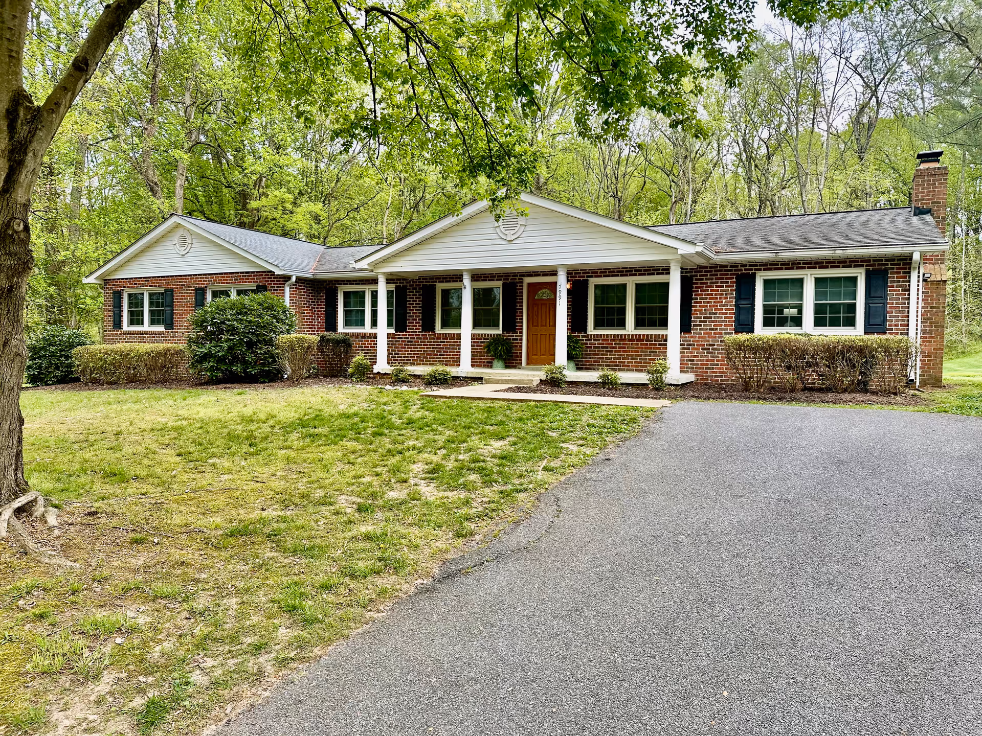 Front exterior view of a single-story brick house with white columns supporting a small porch roof. The house has several windows with dark shutters, a wooden front door, and a chimney on the right side. The driveway leads up to the house, and there is a lawn with some bushes and trees surrounding the property.