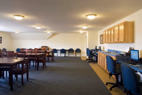 A spacious room with multiple wooden tables and chairs arranged on a carpeted floor. Along one wall, there are several computer workstations with blue office chairs and wooden cabinets mounted above. The room is well-lit with ceiling lights and has a plain white wall in the background with additional chairs lined up against it.