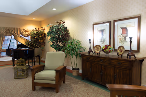 A cozy living room area with a wooden armchair featuring light green cushions, a dark wooden sideboard decorated with two framed pictures, two tall candle holders, and a floral arrangement. In the background, there is a grand piano near a window with draped curtains, and several potted plants adding greenery to the space.