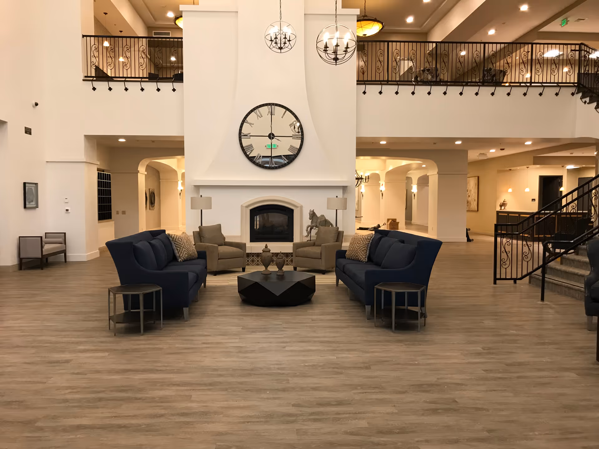 Spacious senior living facility common area with two blue sofas facing each other, two beige armchairs, a black geometric coffee table with decorative items, a large white fireplace with a round clock above it, and a second-floor balcony with wrought iron railing.